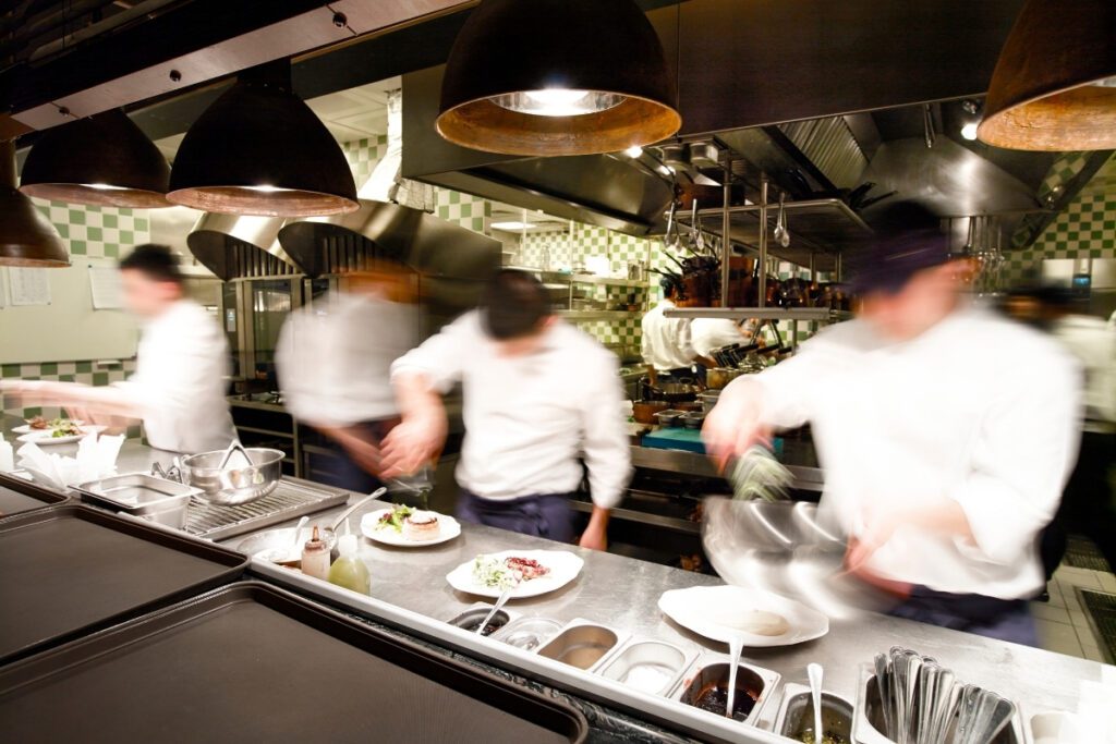 Chefs working at a frantic pace in a high-velocity commercial kitchen, capturing the intense momentum and operational friction of a high-volume hospitality business.